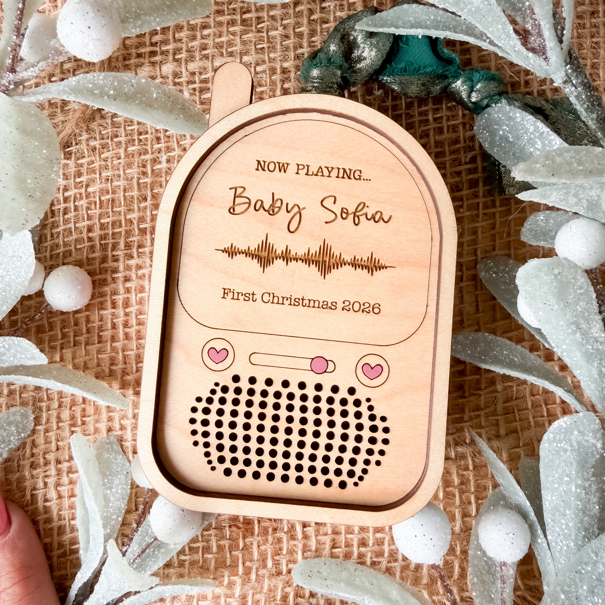 A personalized wooden baby monitor ornament styled on a burlap background with decorative silver leaves and berries. The monitor is engraved with 'NOW PLAYING.