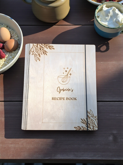 Recipe book with decorative cover on a wooden table with food items.