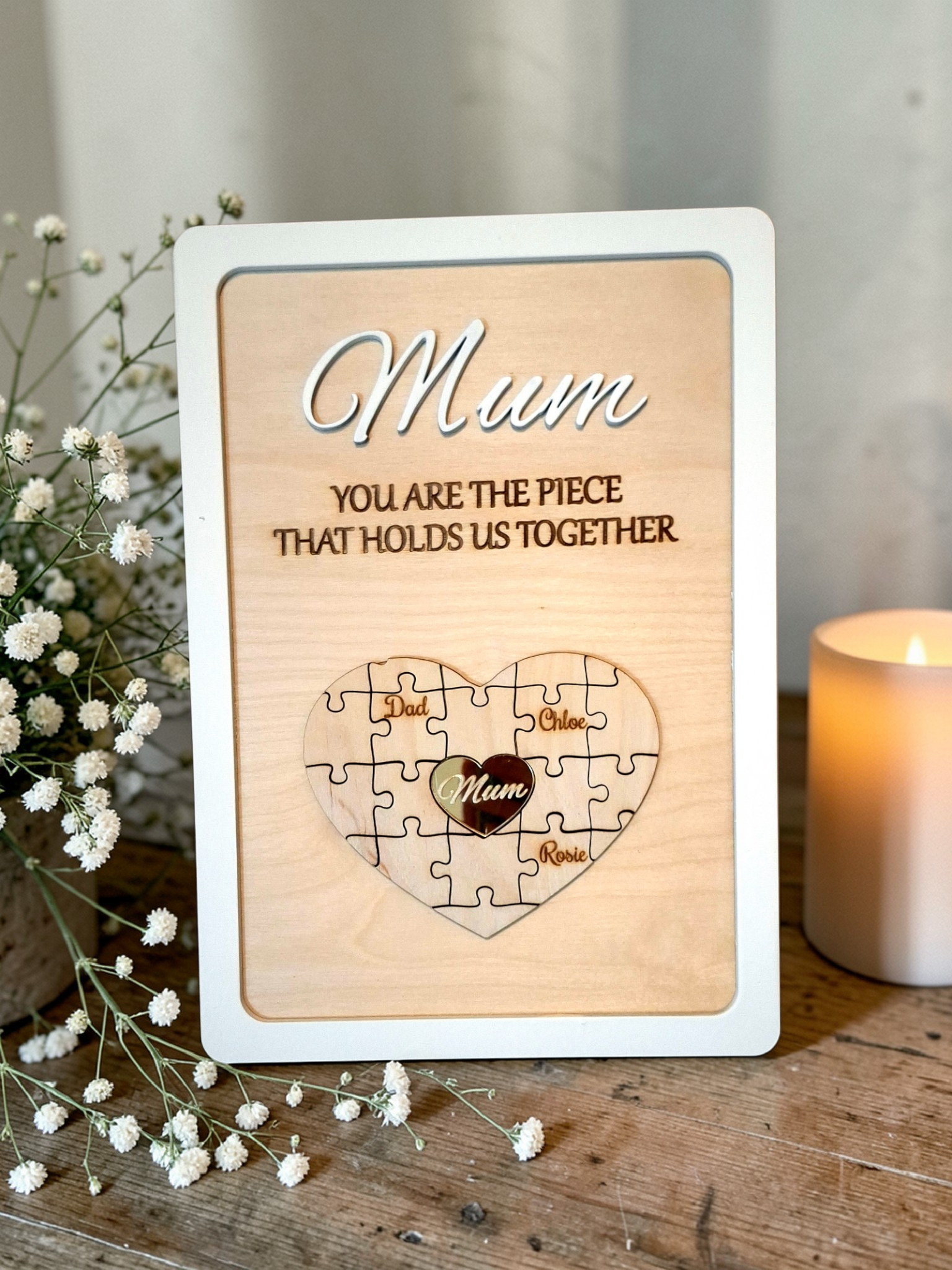 Wooden plaque with 'Mum, you are the piece that holds us together' text and a puzzle heart design on a wooden surface with flowers and a candle.