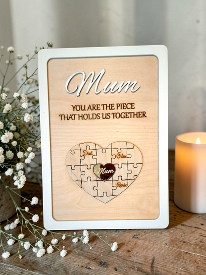 Wooden plaque with 'Mum, you are the piece that holds us together' text and a puzzle heart design on a wooden surface with flowers and a candle.