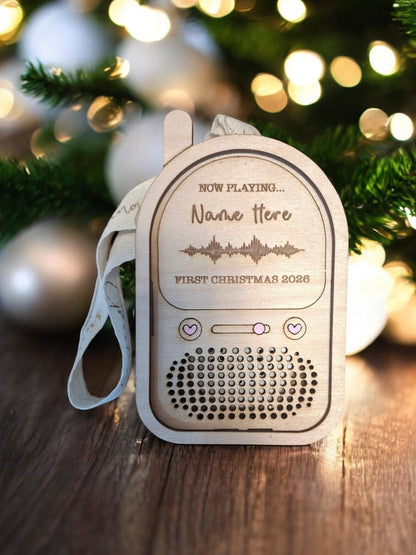 A personalized wooden first birthday Christmas speaker with a white fabric strap, resting on a wooden surface with a blurred Christmas tree in the background.
