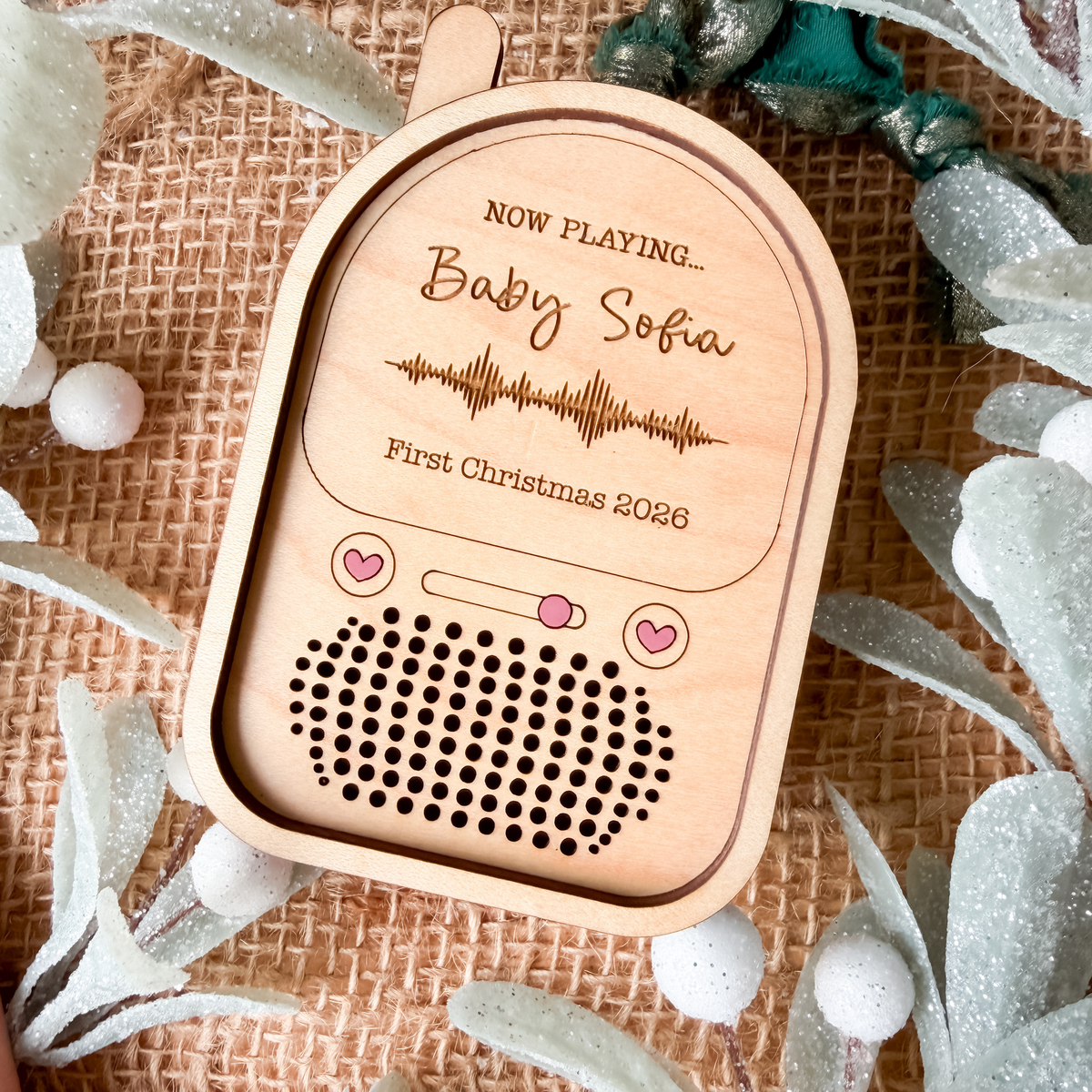 A personalized wooden baby monitor ornament styled on a burlap background with decorative greenery. The ornament is engraved with 'NOW PLAYING.