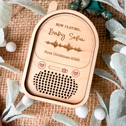 A personalized wooden baby monitor ornament styled on a burlap background with decorative greenery. The ornament is engraved with 'NOW PLAYING.